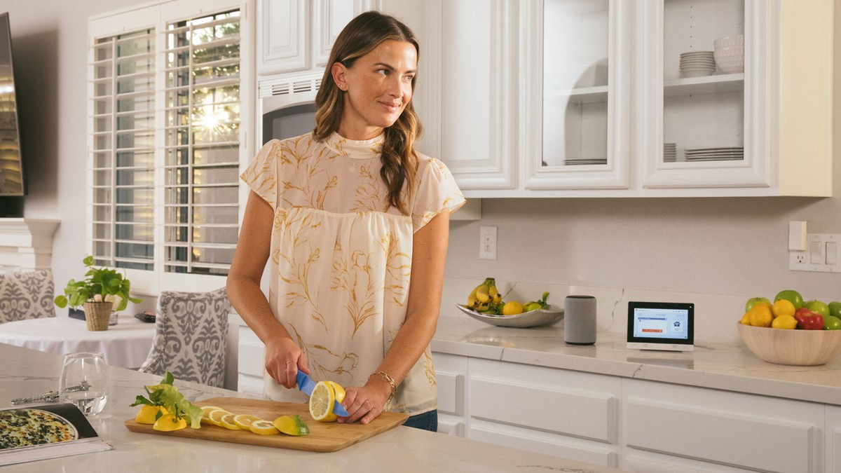 A woman slicing lemons in a modern kitchen with white cabinets and a marble countertop, accompanied by a smart home control tablet and speaker in the background.